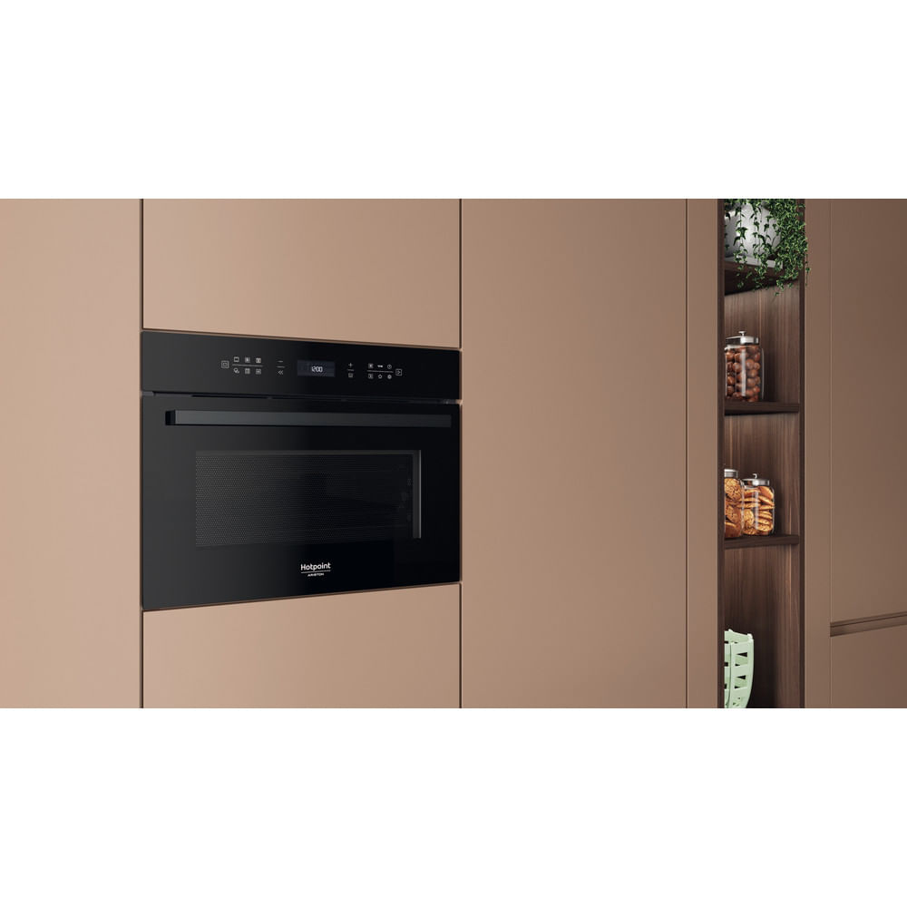 Un forno a microonde nero incorporato con orologio digitale è installato in un mobile da cucina marrone. Scaffali contengono barattoli e una pianta.