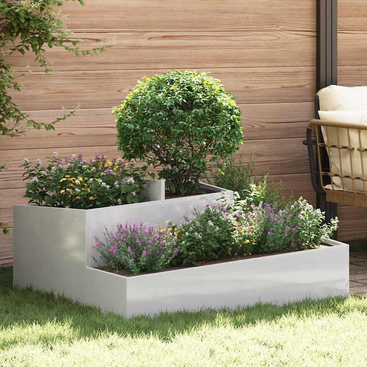 Una fioriera bianca a più livelli con fiori e un cespuglio. Una recinzione in legno e una sedia sullo sfondo. Erba verde in primo piano.