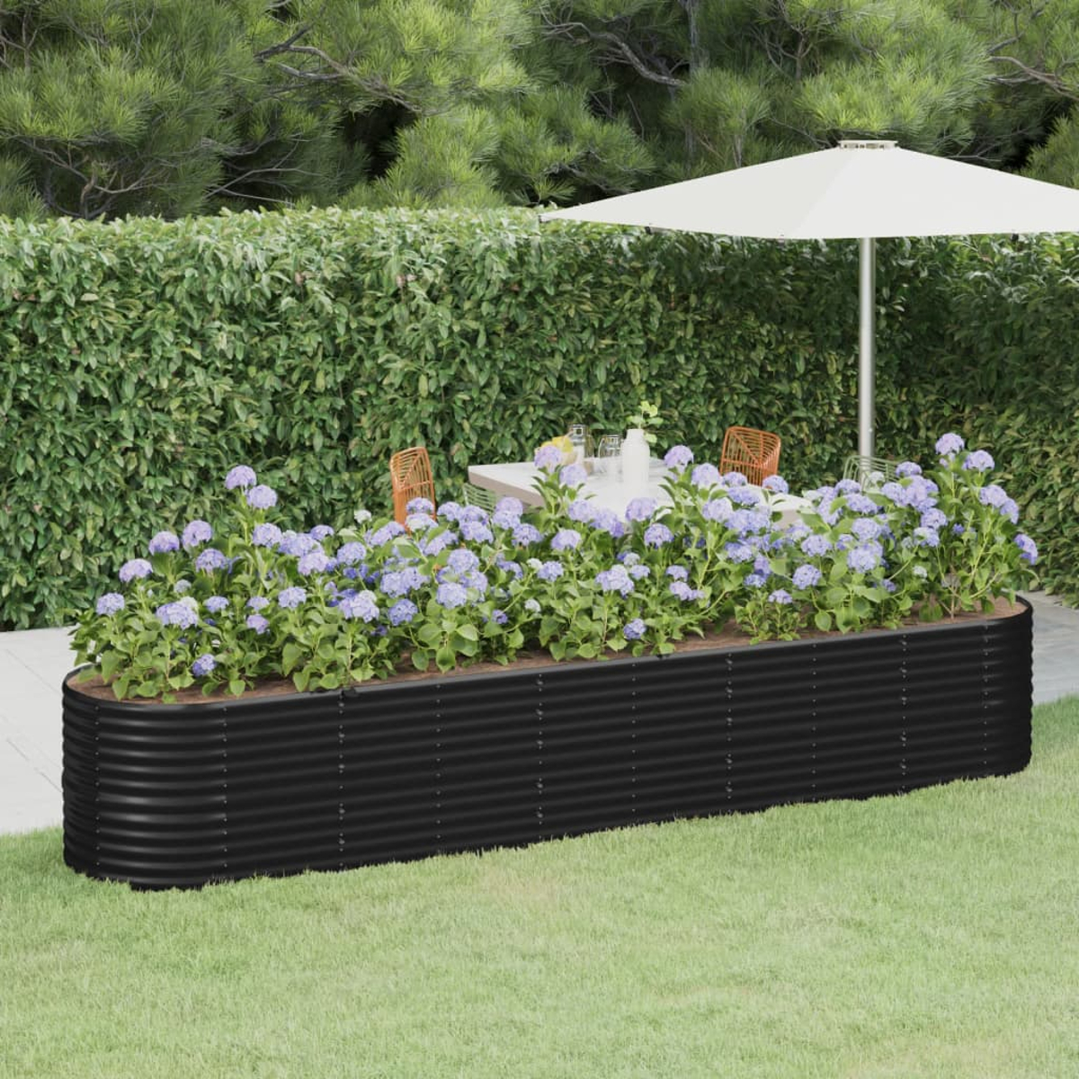 Ein schwarzes Hochbeet mit blauen Blumen steht auf Gras. Ein weißer Schirm beschattet einen Tisch mit Stühlen. Grüne Hecke im Hintergrund.