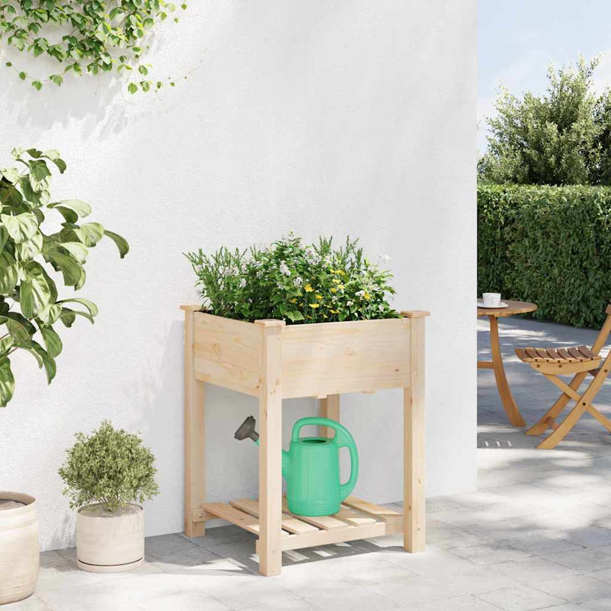 Fioriera in legno con fiori, annaffiatoio verde e alberello. Sfondo muro bianco con verde e sedute all'aperto.