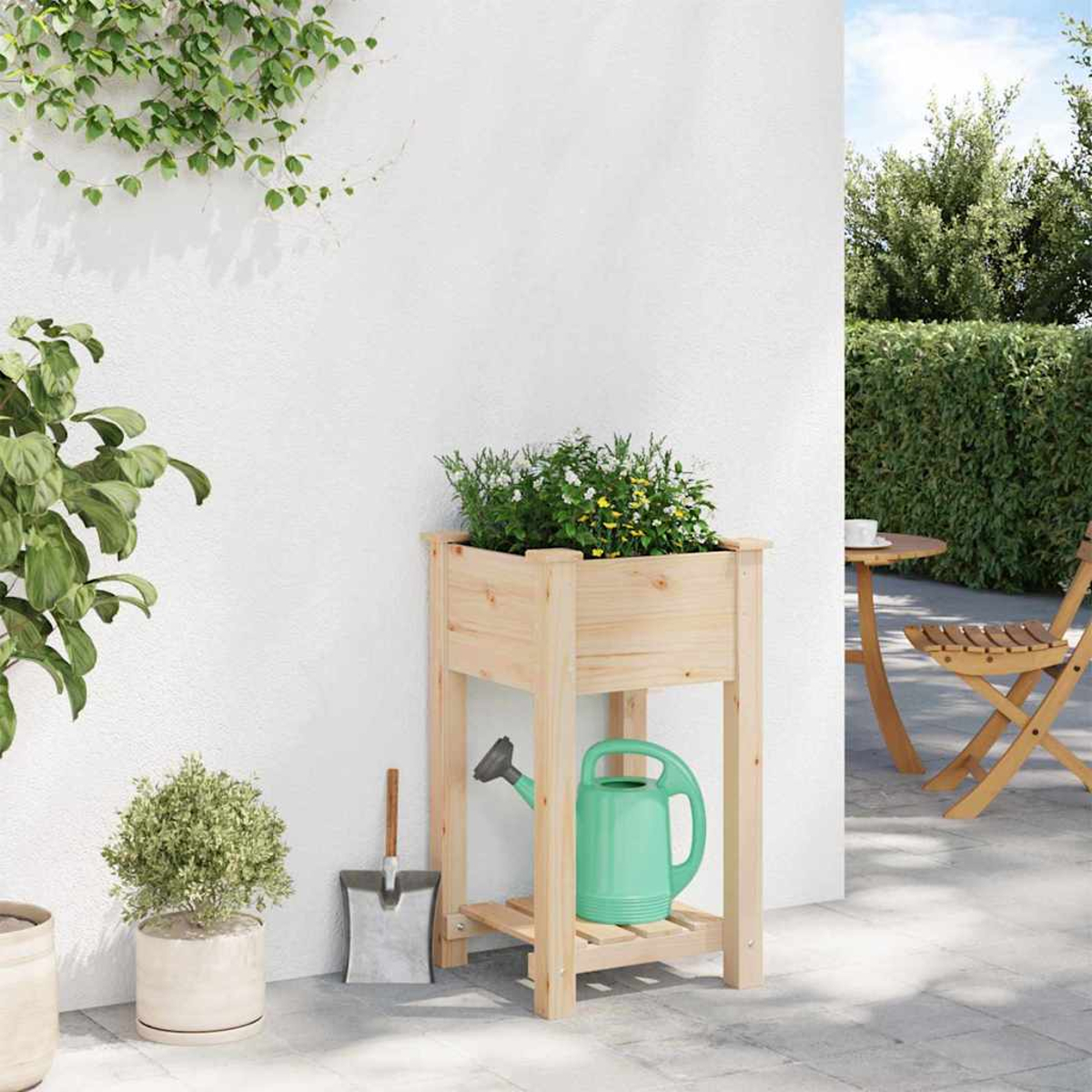 Una fioriera in legno con fiori, un annaffiatoio verde e una pala sono appoggiati a un muro bianco. Un tavolo e una sedia sono sullo sfondo.