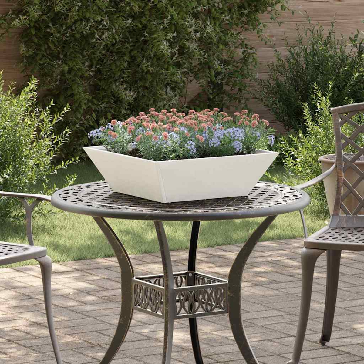 Una fioriera bianca su un tavolo di metallo contiene fiori rosa e blu. Il tavolo è su un patio di mattoni. Cespugli verdi sullo sfondo.