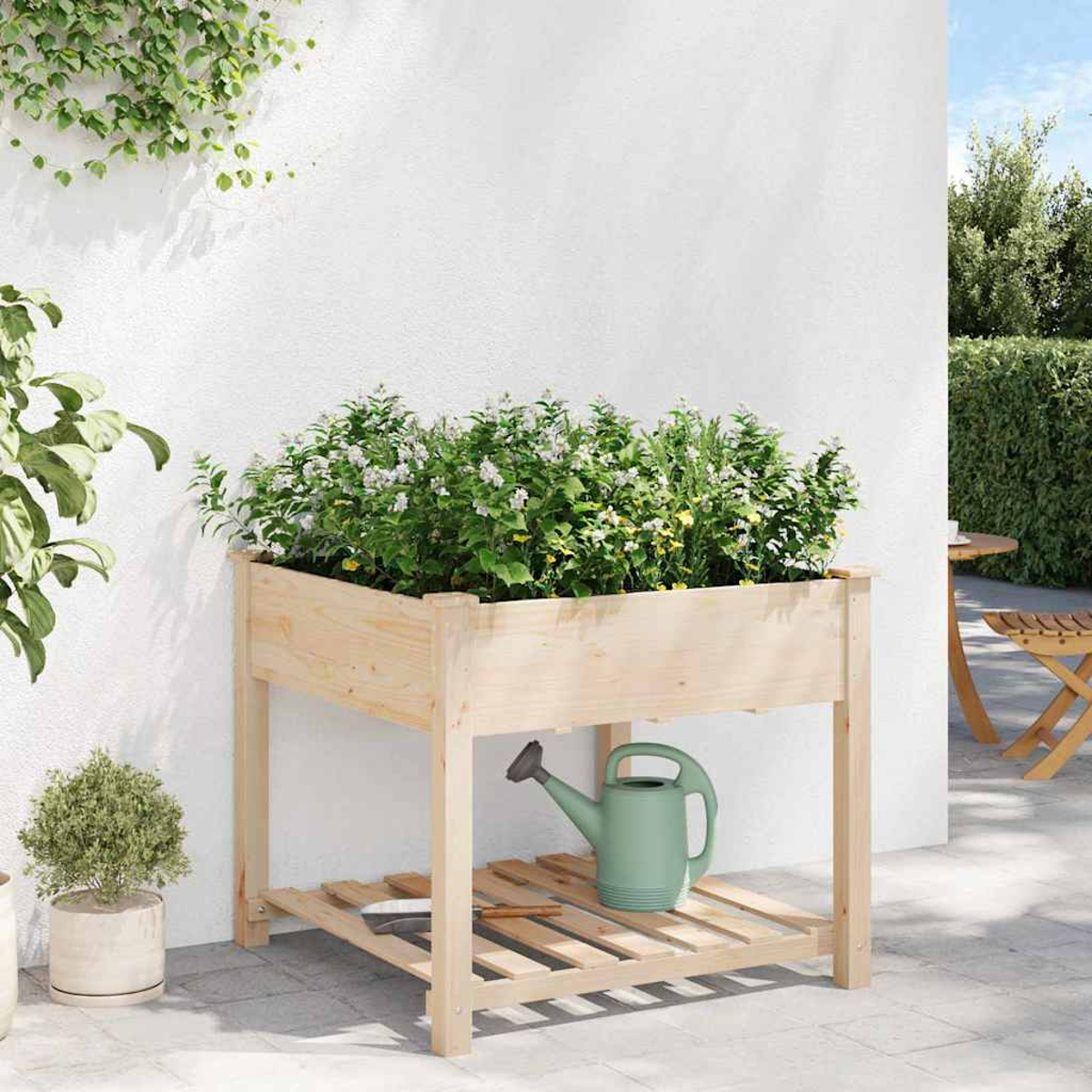 Fioriera in legno con piante. Un annaffiatoio verde sotto. Piante verdi sullo sfondo, muro bianco e un tavolo sono visibili.