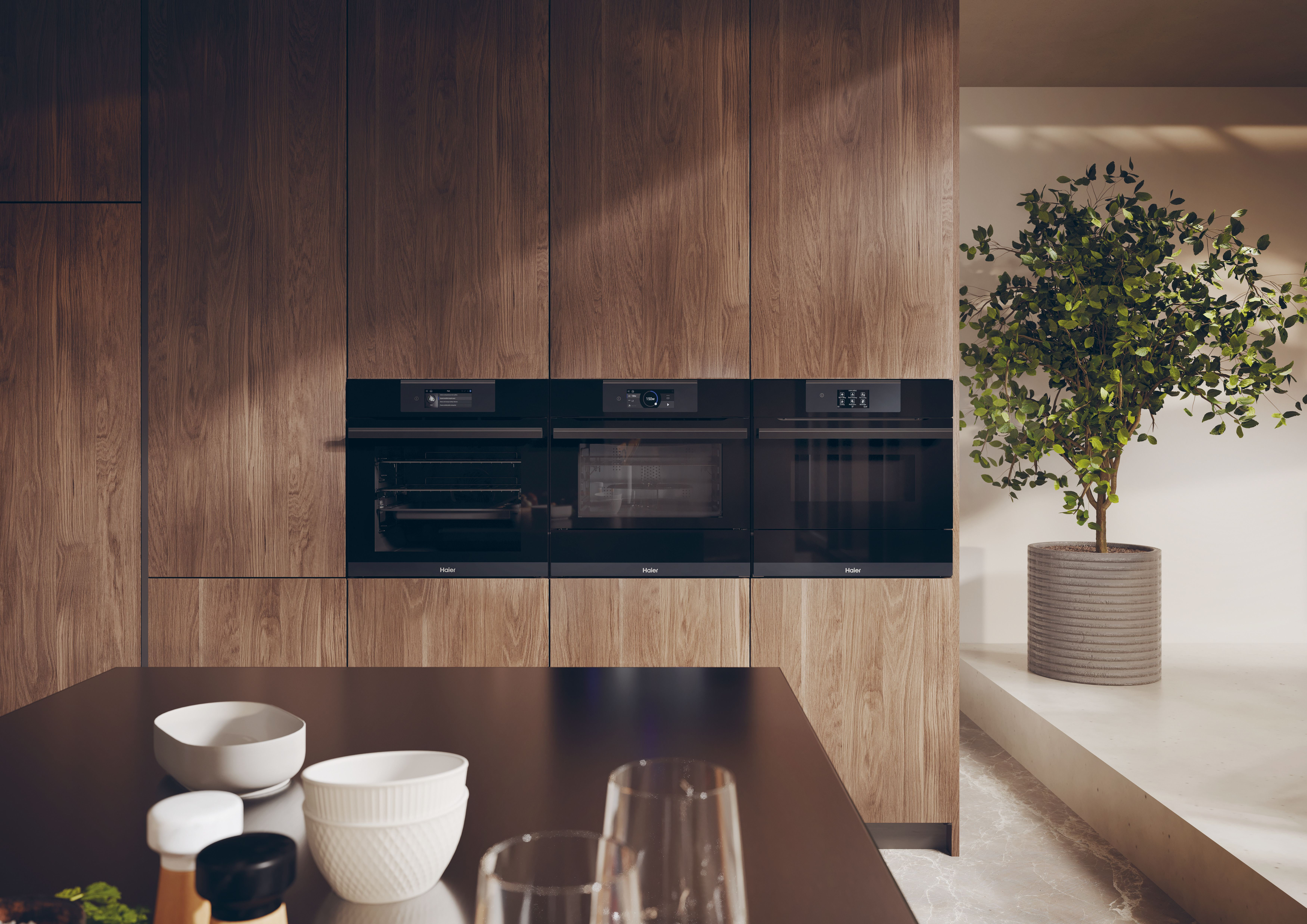 Cucina con tre forni neri, armadi in legno, pianta in vaso e piano scuro con ciotole e bicchieri.