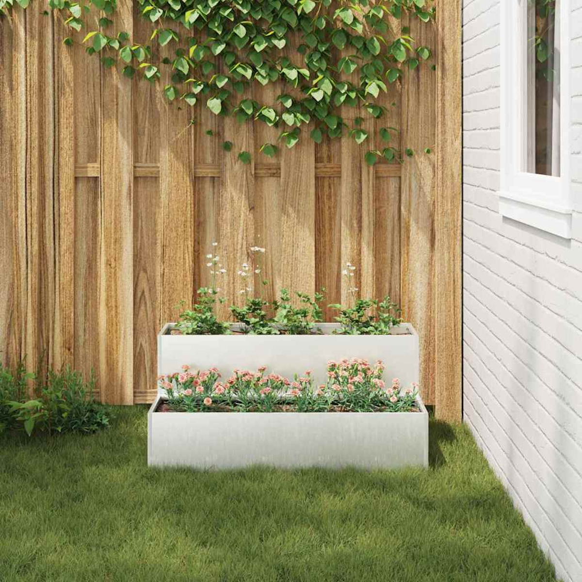 Fioriera a due livelli con piante rosa e verdi sull'erba. Recinzione in legno e muro di mattoni sullo sfondo, viti sulla recinzione.