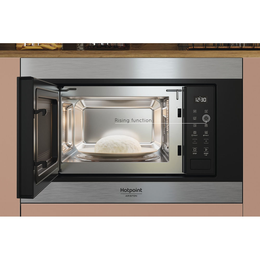Een magnetron met een open deur. Binnenin een bord met rijzend deeg. De oven heeft een zwart bedieningspaneel. Hotpoint Ariston-logo.