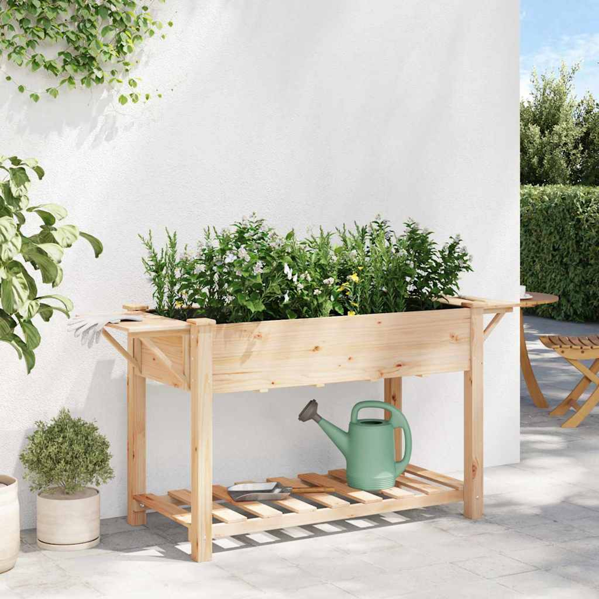 Fioriera in legno con piante, annaffiatoio e attrezzi. Contro un muro bianco, con verde e un tavolo sullo sfondo.