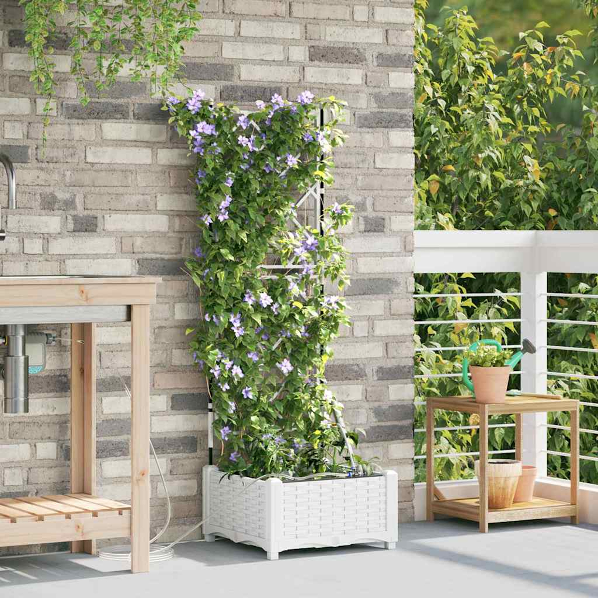 Ein weißer Pflanzkübel mit Kletterblumen vor einer Ziegelmauer. Ein kleiner Holztisch mit Töpfen und einer Gießkanne steht daneben.