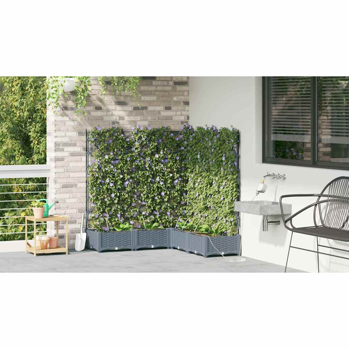 Una scena di patio mostra un muro di mattoni con piante, una siepe verde con fiori viola e fioriere grigie. A destra una sedia e un lavandino.