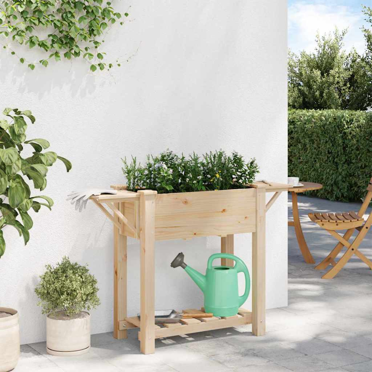 Fioriera in legno con piante, annaffiatoio e attrezzi da giardinaggio. Muro bianco con verde. Tavolo e sedia sullo sfondo.