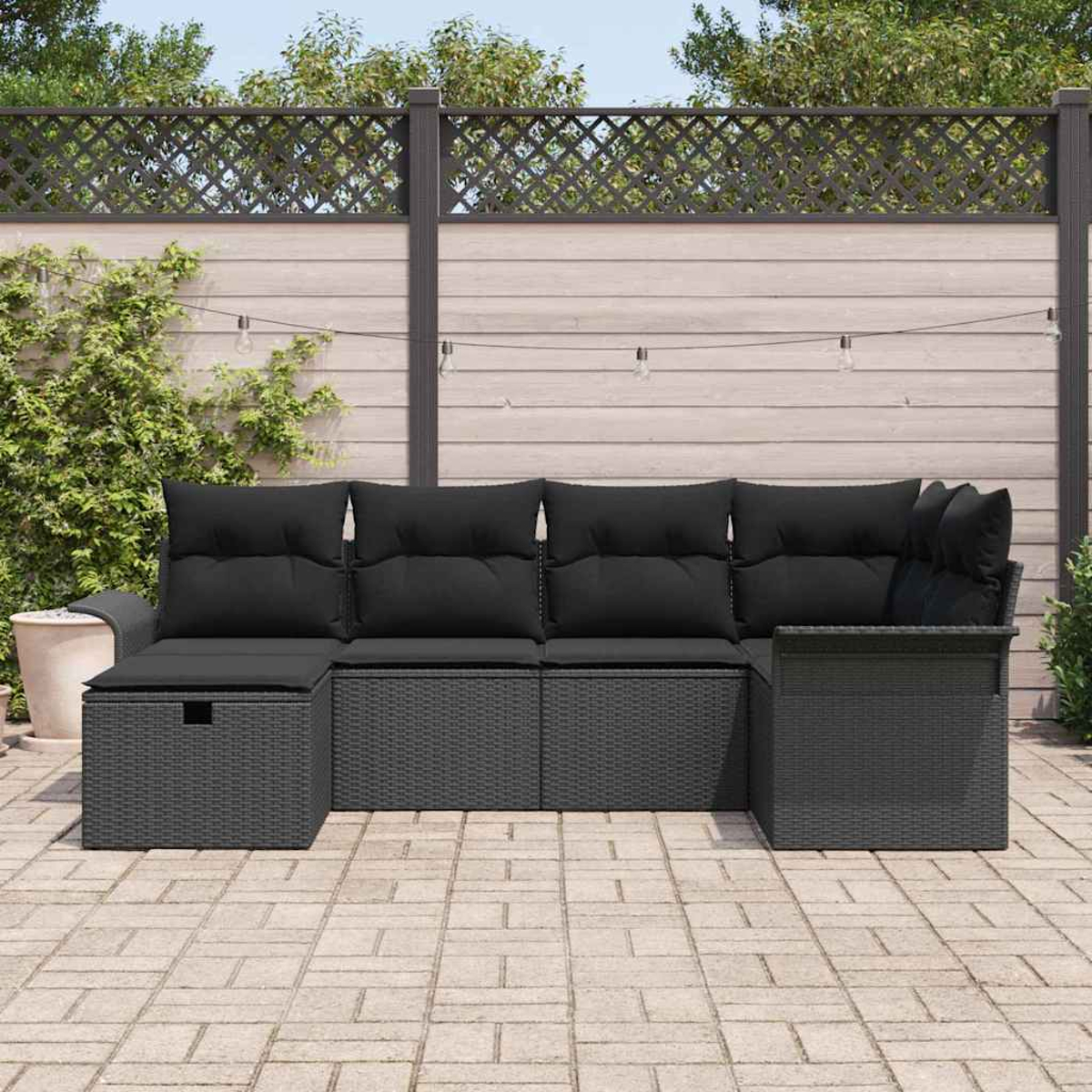 Graues Rattan-Outdoor-Ecksofa mit schwarzen Kissen auf einer Ziegelterrasse, mit Zaun und Pflanzen im Hintergrund.