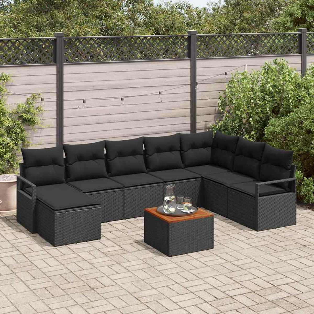 Ein schwarzes Ecksofa aus Korbgeflecht mit schwarzen Kissen steht auf einer Terrasse. Davor steht ein kleiner Tisch mit einem Glaskrug.