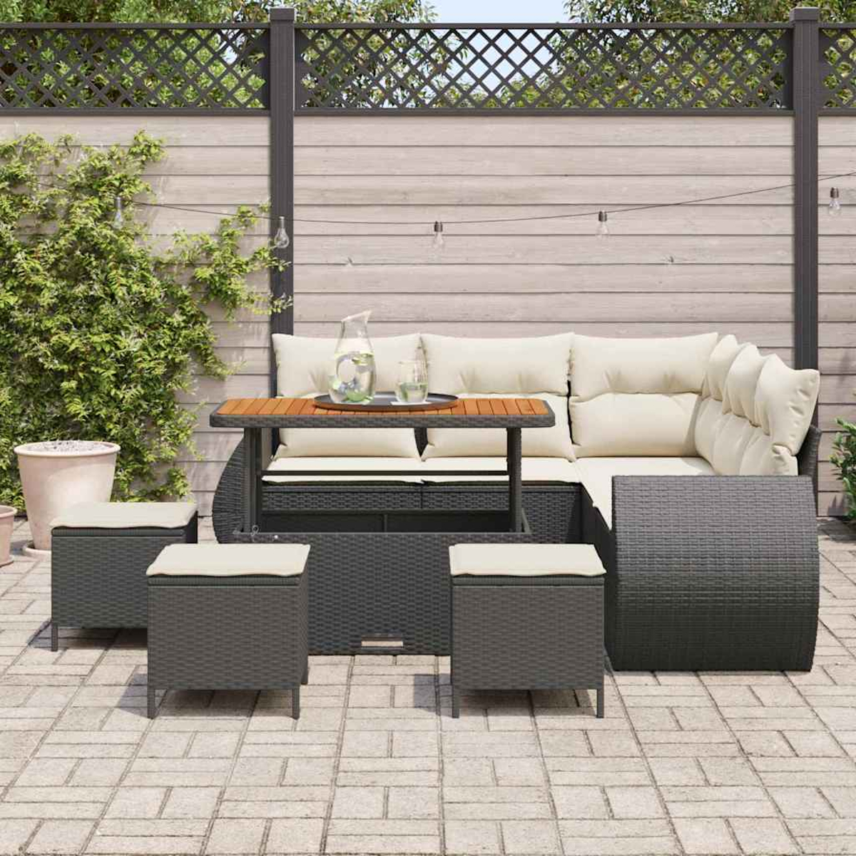 Terrassenset mit dunkelgrauen Korbmöbeln, beigen Kissen und einer Holzplatte. Enthält Sofa, Tisch und Hocker, auf Pflastersteinen.