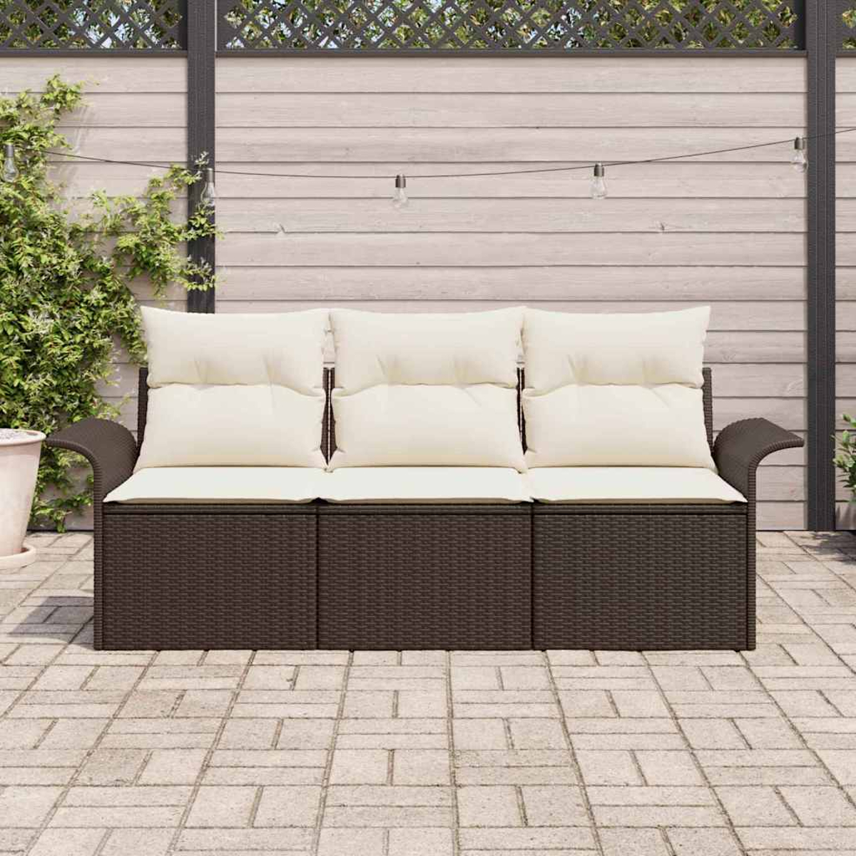 Ein braunes Outdoor-Sofa aus Korbgeflecht mit cremefarbenen Kissen steht auf einer Backstein-Terrasse. Im Hintergrund ein Holzzaun und Lichterketten.