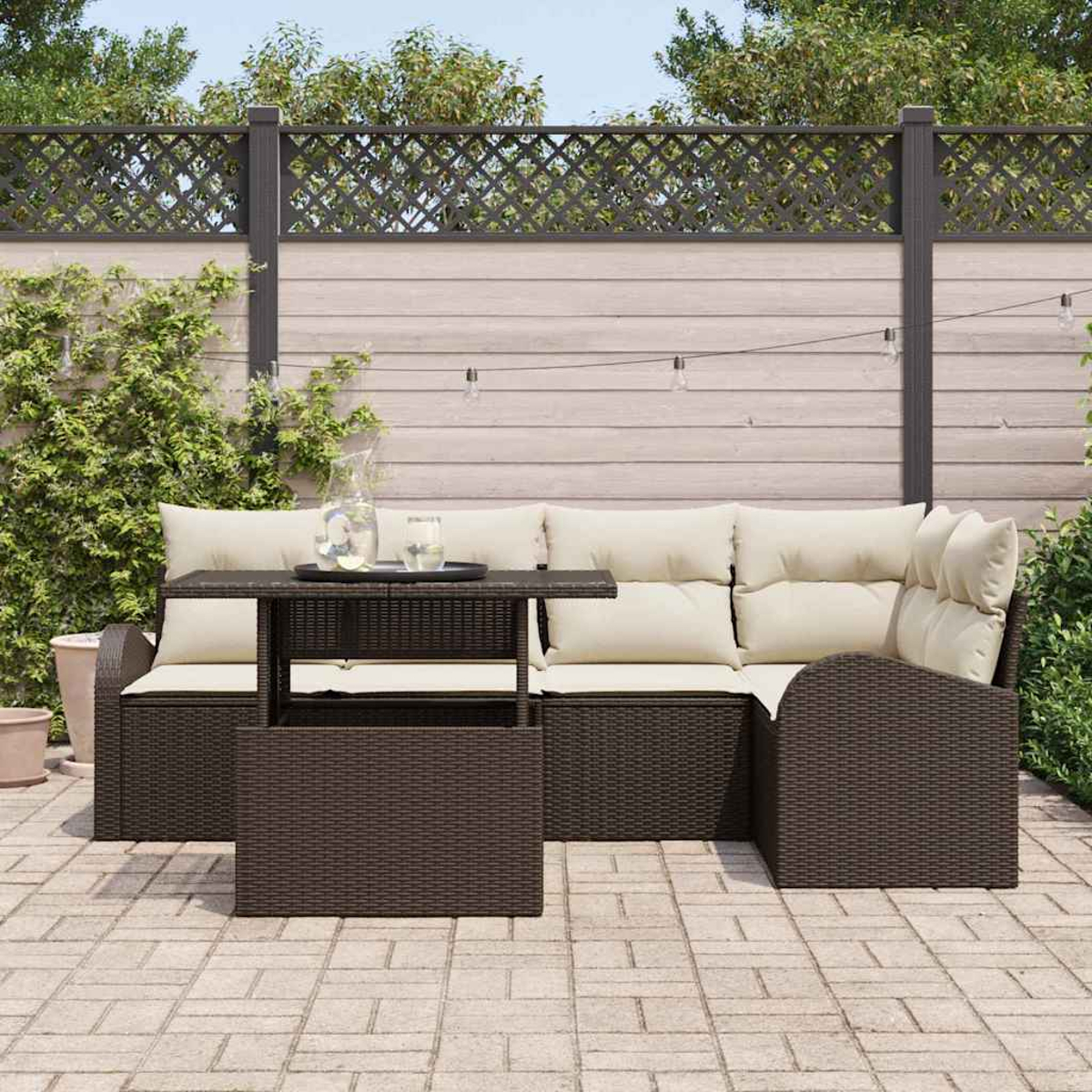 Gartenlounge mit Tisch. Beige Kissen auf braunen Möbeln. Holzzaun im Hintergrund mit Pflanzen und Himmel.