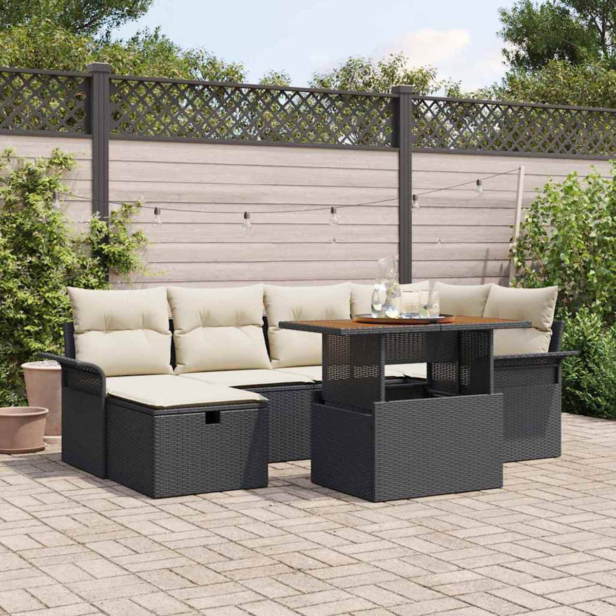 Gartenmöbel-Set mit Sofa, Tisch und Kissen. Auf einer gepflasterten Terrasse mit Zaun und Grün im Hintergrund.