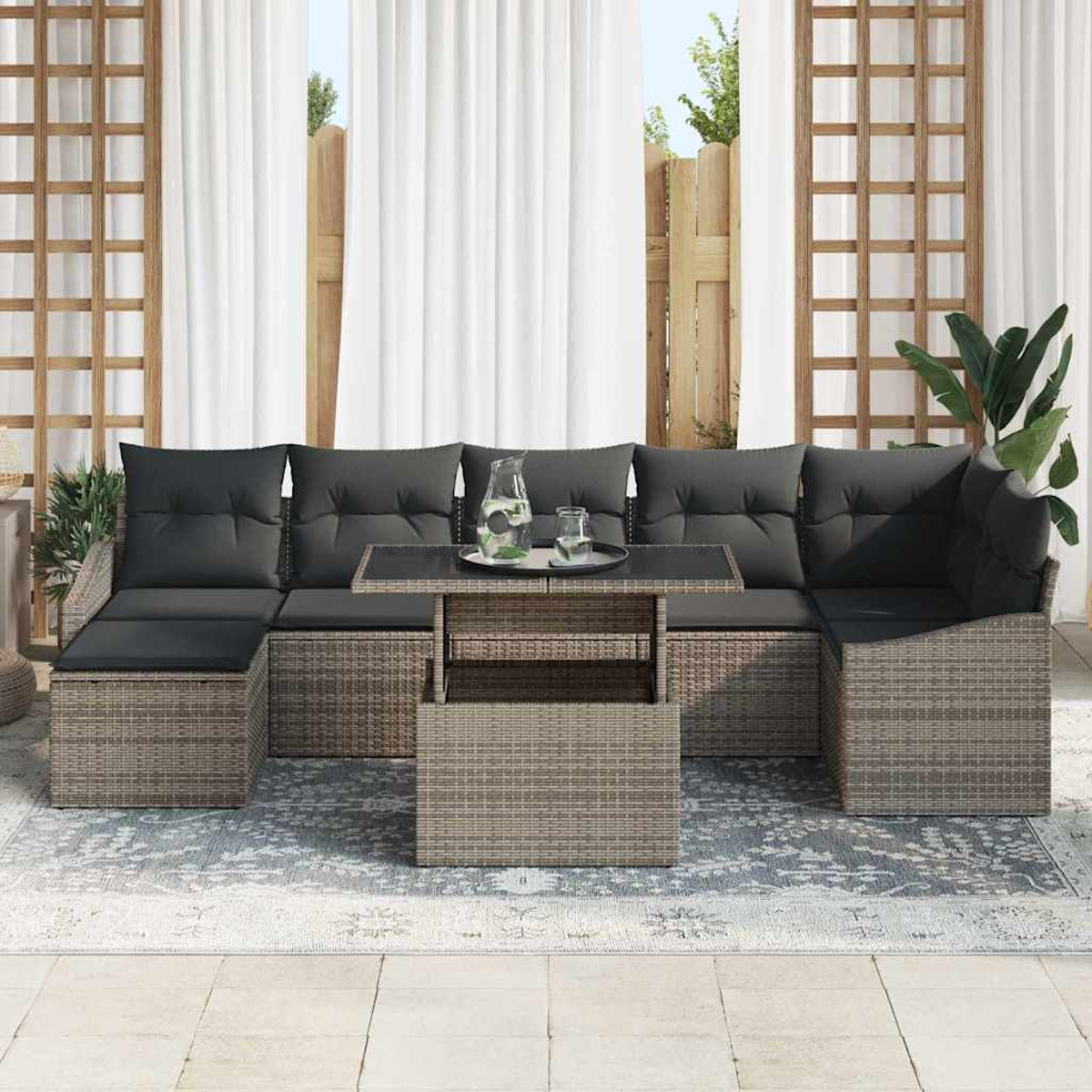 Outdoor-Ecksofa mit dunklen Kissen und Korbgeflecht-Basis. Ein Couchtisch mit Glasplatte und eine Glaskaraffe mit Gläsern darauf.