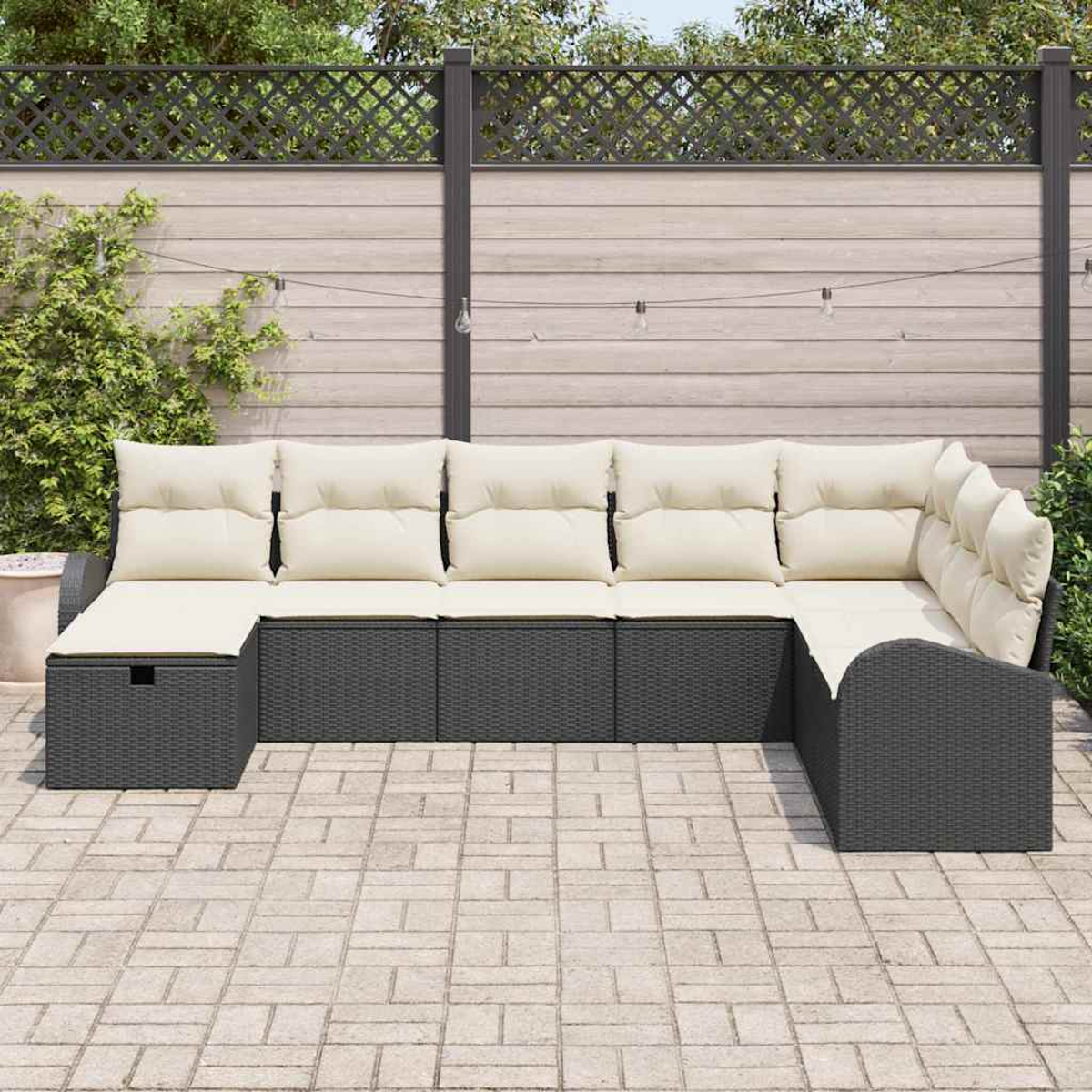 Ein Outdoor-Ecksofa mit cremefarbenen Kissen steht auf einer Backstein-Terrasse. Im Hintergrund befindet sich ein Holzzaun mit Lichterketten.