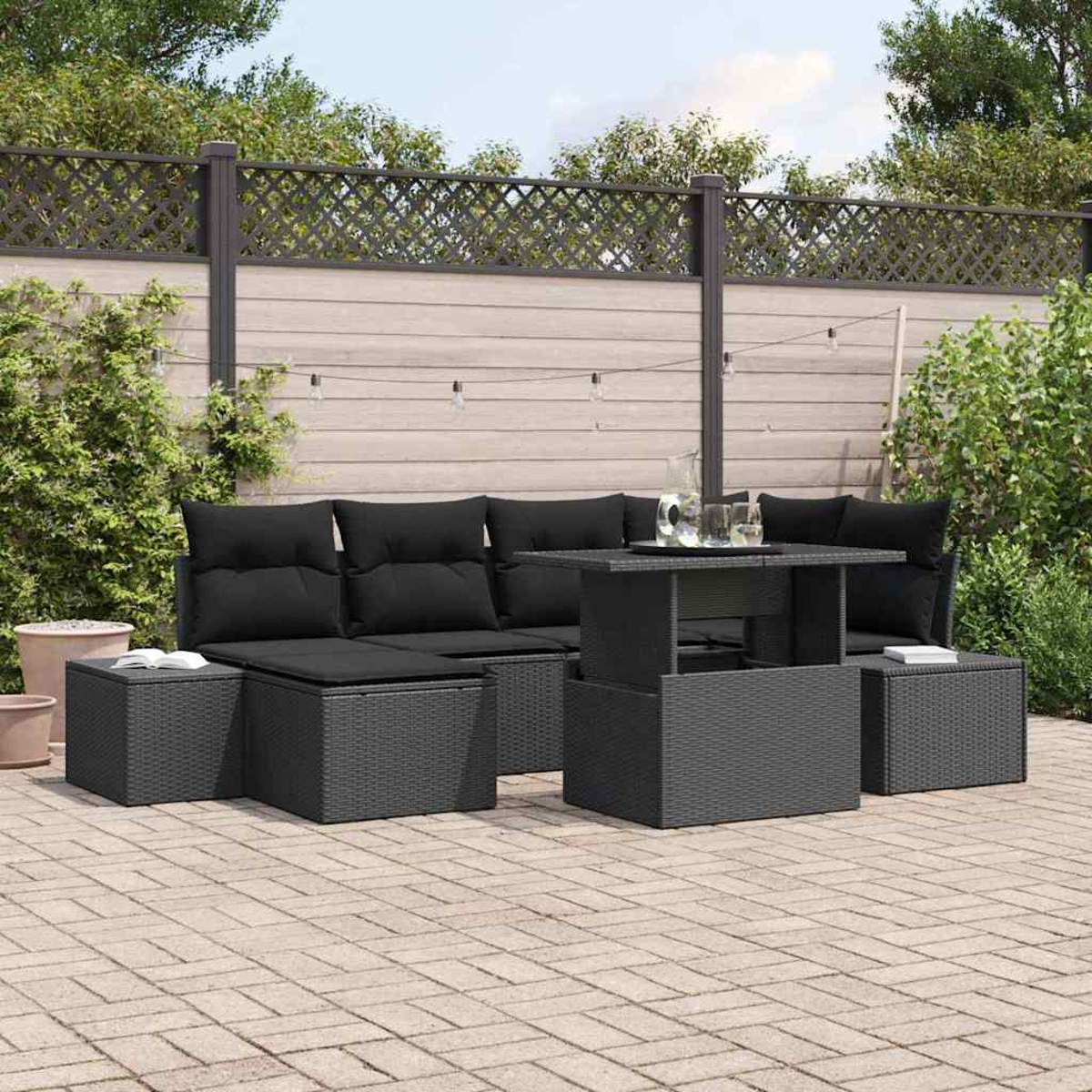 Schwarzes Outdoor-Geflechtmöbel-Set mit Kissen, einem Tisch und Ottomanen auf einer Terrasse. Im Hintergrund ein Zaun und Grün.