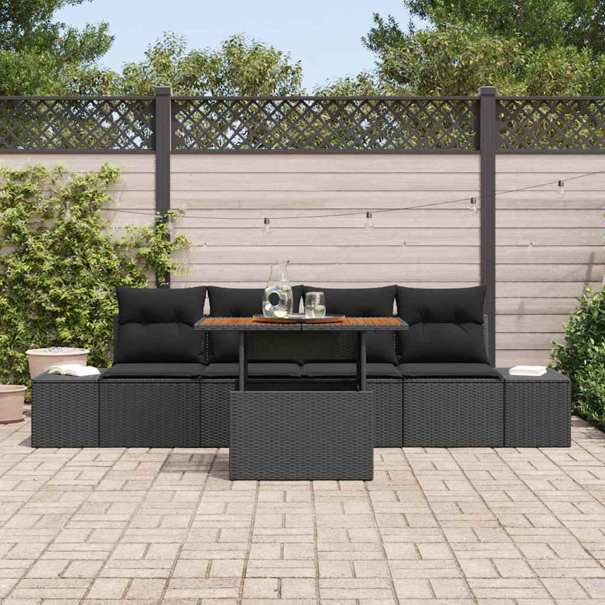 Garten-Terrassenset mit schwarzen Kissen, einem Holztisch und einer grauen Korbbasis. Im Hintergrund sind ein Zaun und Grün zu sehen.