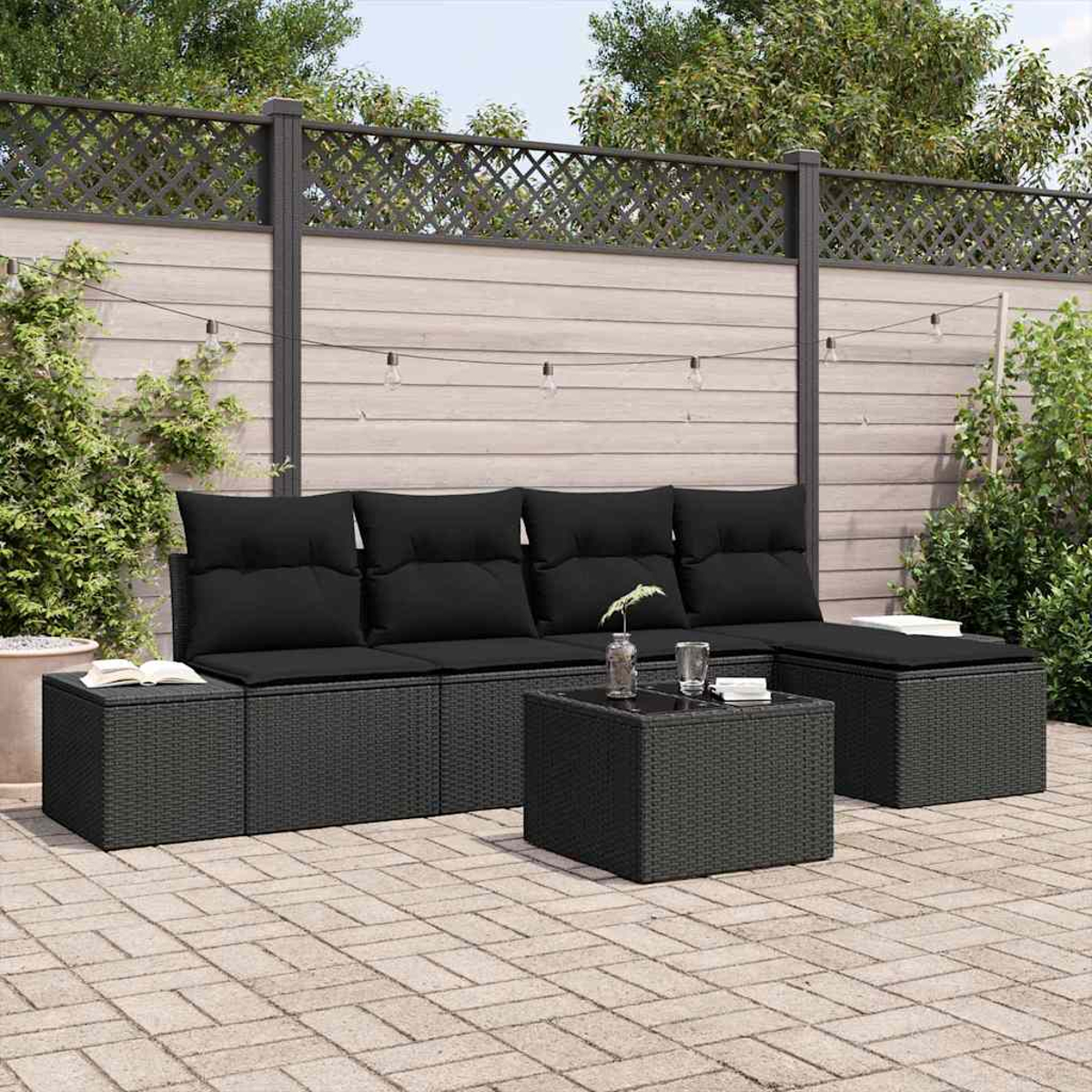 Schwarzes Outdoor-Korbssofa mit schwarzen Kissen, Beistelltischen und einem Couchtisch auf einer Backsteinterrasse. Im Hintergrund ein Holzzaun.