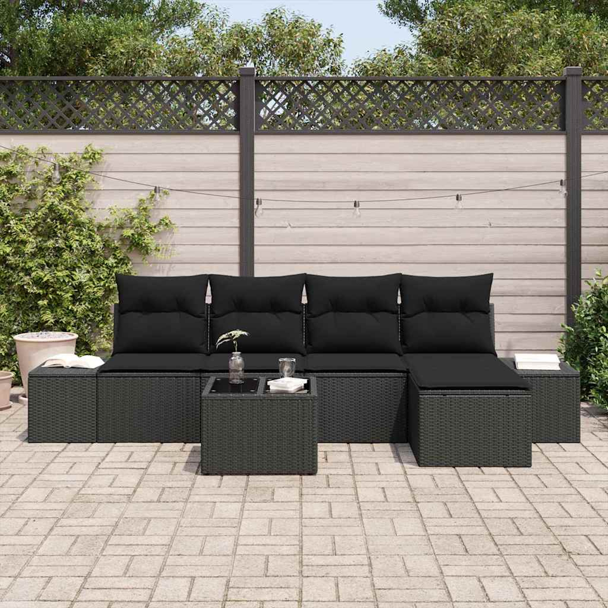 Garten-Set mit schwarzen Kissen auf grauen Korbmöbeln, kleiner Tisch mit Glas und Pflanzen, vor einem Zaunhintergrund.