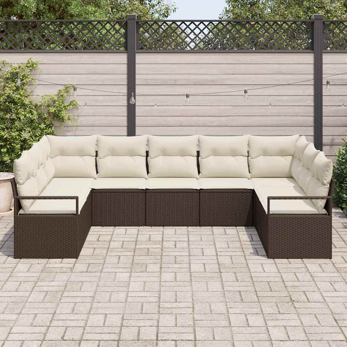 Ein U-förmiges Outdoor-Sofa mit cremefarbenen Kissen auf einem braunen Korbrahmen. Das Sofa steht auf einer Ziegelterrasse mit Holzzaun im Hintergrund.