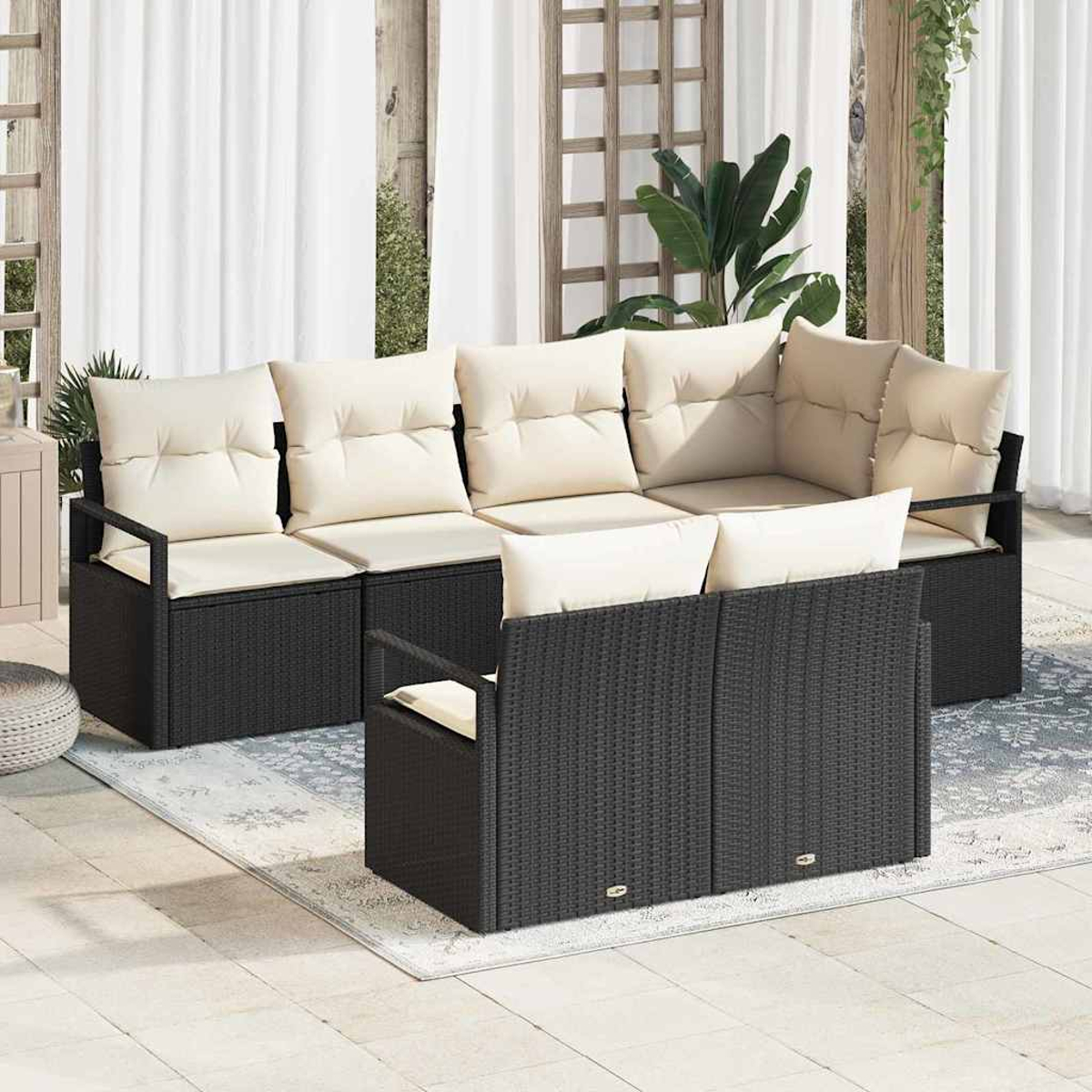 Sofá seccional de mimbre negro para exteriores con cojines crema. Colocado sobre una alfombra de patio, con cortinas y vegetación de fondo.