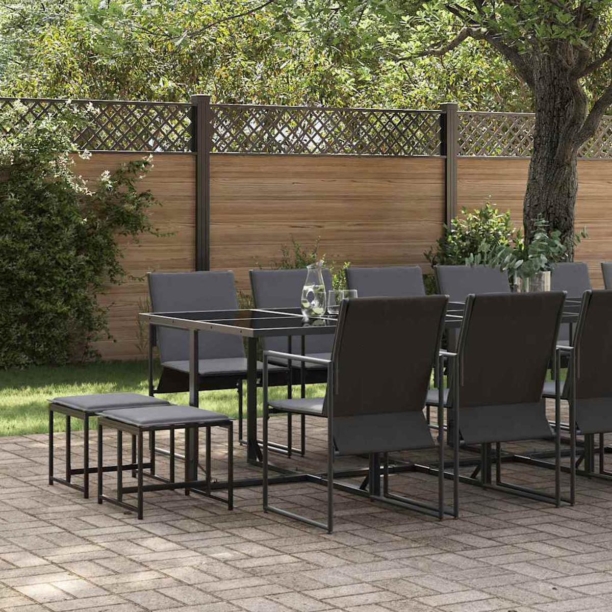 Essgruppe im Freien: Tisch mit Glasplatte, graue Stühle, Bänke auf einer Backstein-Terrasse. Hintergrund ist ein Holzzaun und Bäume.