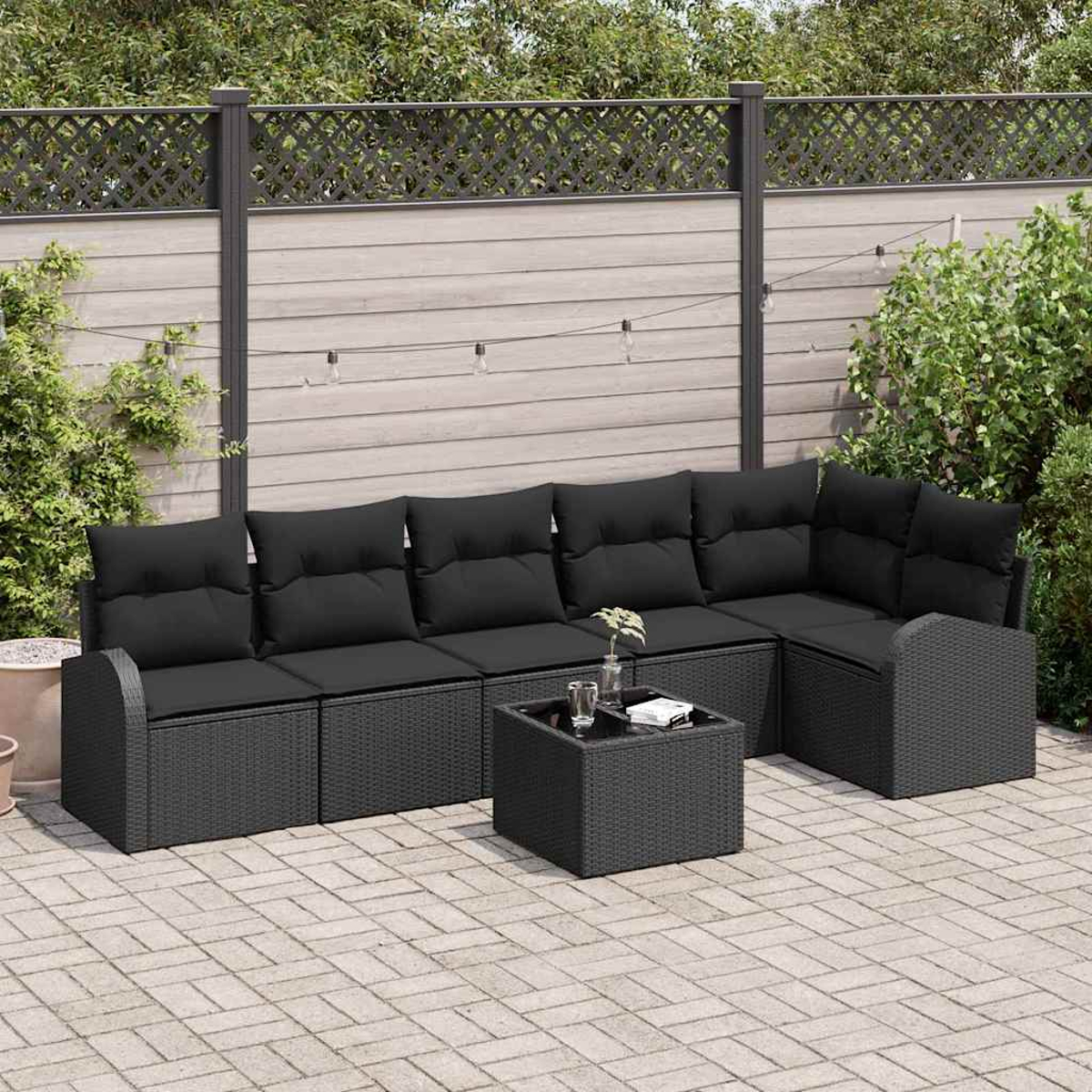 Sofá de mimbre negro para exteriores con cojines negros. Una mesa cuadrada con tapa de cristal y una planta pequeña. Fondo con valla y plantas.