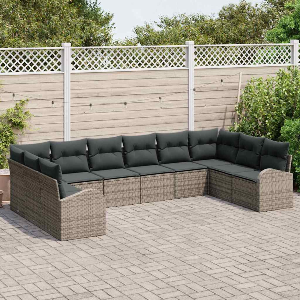 Ein graues Korbsessel-Ecksofa mit dunkelgrauen Kissen steht auf einer Ziegelsteinterrasse. Ein Holzzaun und grüne Pflanzen sind im Hintergrund.