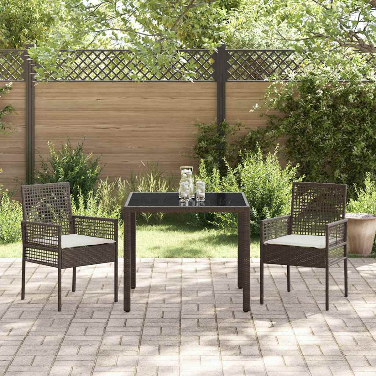 Juego de patio con dos sillas y una mesa. El juego es marrón y tiene cojines blancos. El fondo tiene una valla y plantas.