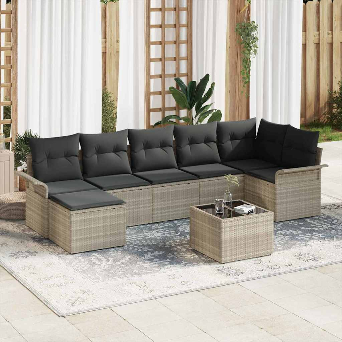 Ein graues Outdoor-Sofa mit schwarzen Kissen steht auf einem gemusterten Teppich, davor ein kleiner Tisch. Weiße Vorhänge und ein Holzzaun im Hintergrund.