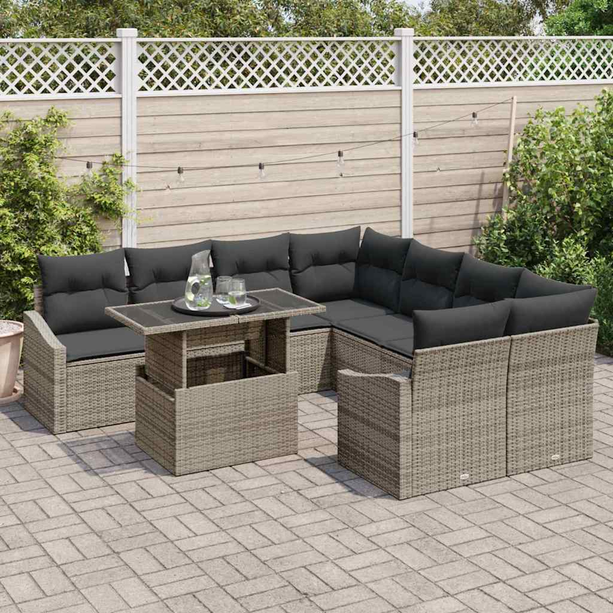 Garten-Terrassenset mit grauen Korbmöbeln, schwarzen Kissen und einem Glastisch. Im Hintergrund ein weißer Zaun und grüne Büsche.