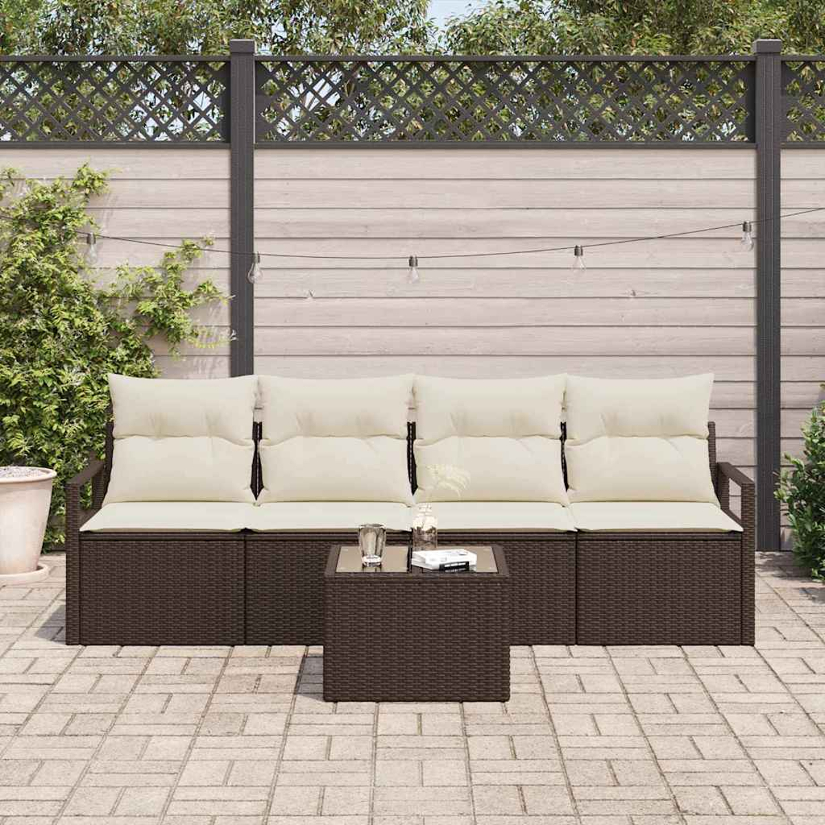 Garten-Set: braunes Korbmöbel mit cremefarbenen Kissen, kleiner Tisch und Backstein-Terrasse. Zaun und Pflanzen im Hintergrund.