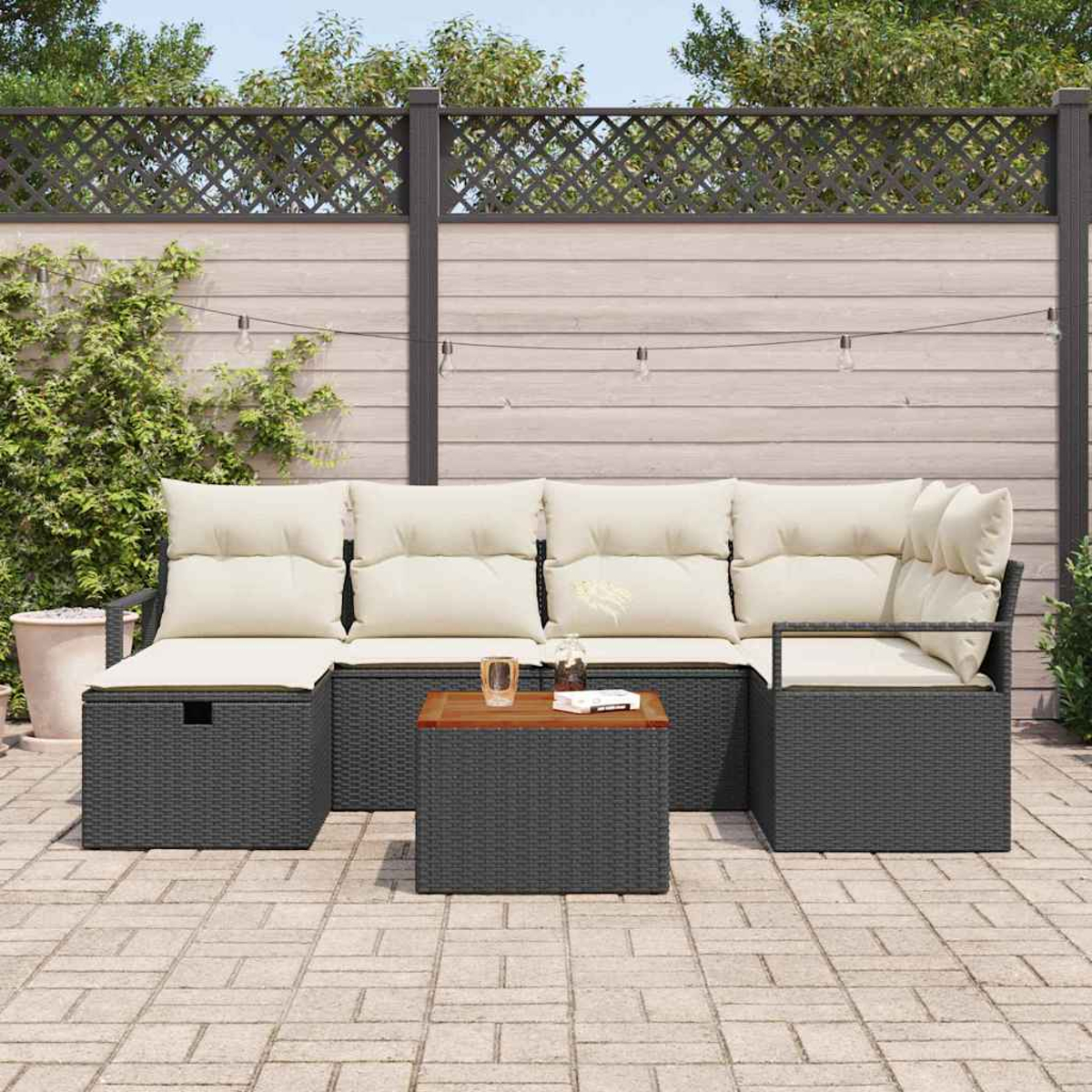 Juego de sofás de mimbre para exteriores con cojines beige, mesa de madera y luces de cadena en un patio con valla al fondo.