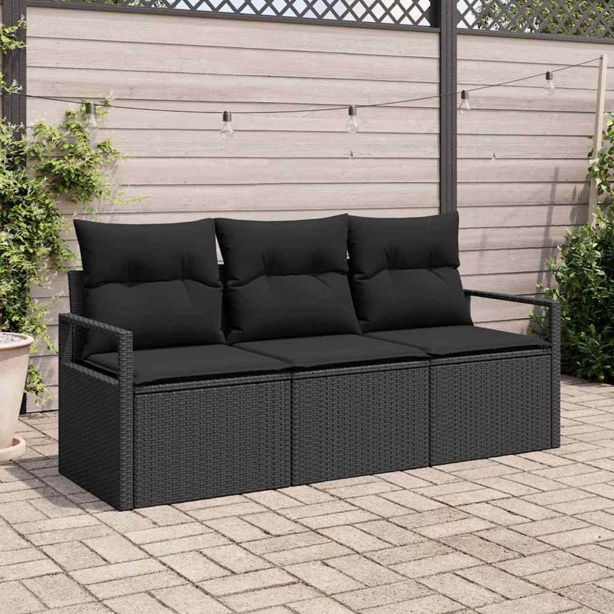Schwarzes Outdoor-Sofa aus Korbgeflecht mit schwarzen Kissen. Der Hintergrund hat einen Holzzaun und Lichterketten. Gepflasterte Terrasse.