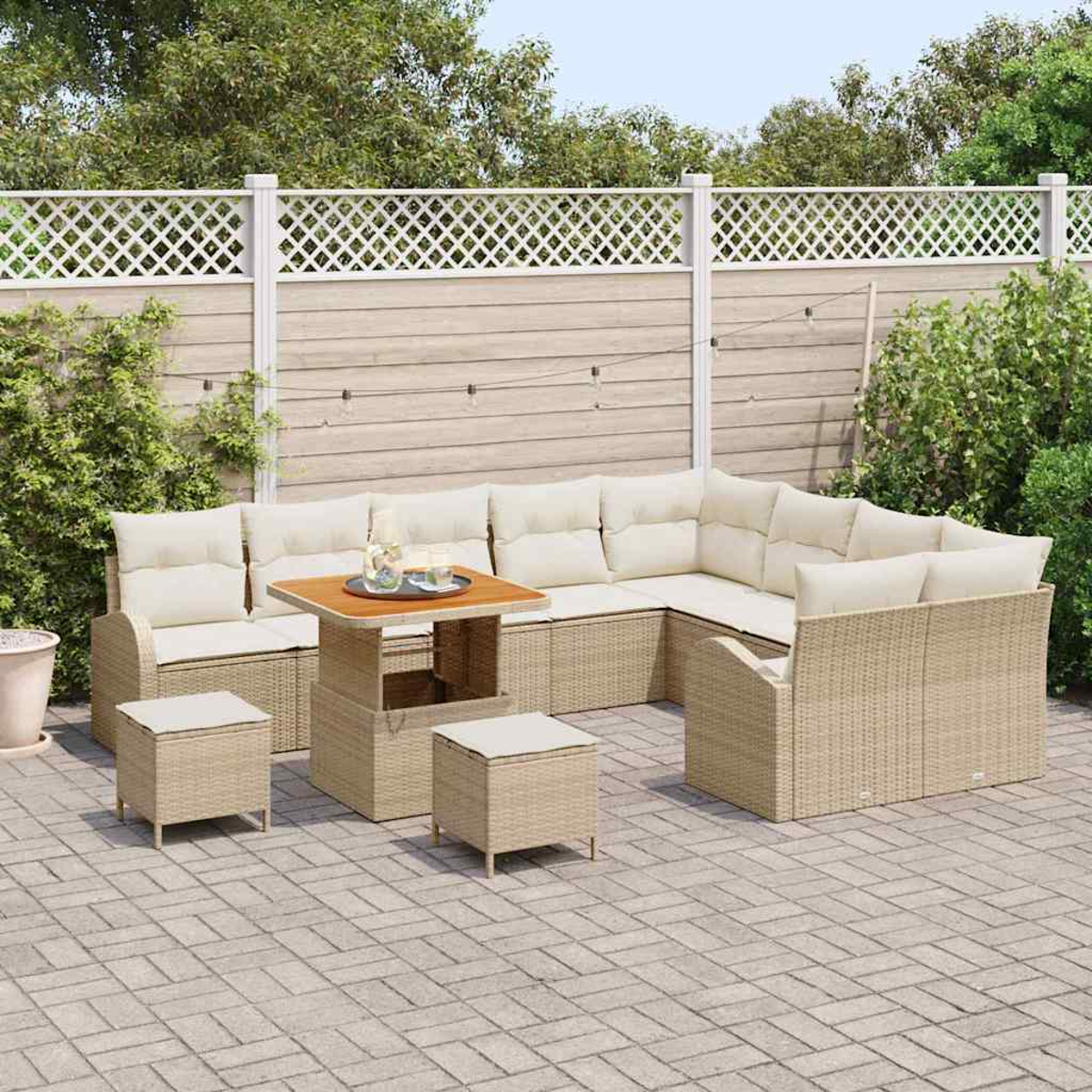 Gartenmöbel-Set aus Korbgeflecht: Ecksofa, quadratischer Tisch, zwei Hocker, cremefarbene Kissen, auf Terrasse, mit Zaun und Pflanzen im Hintergrund.