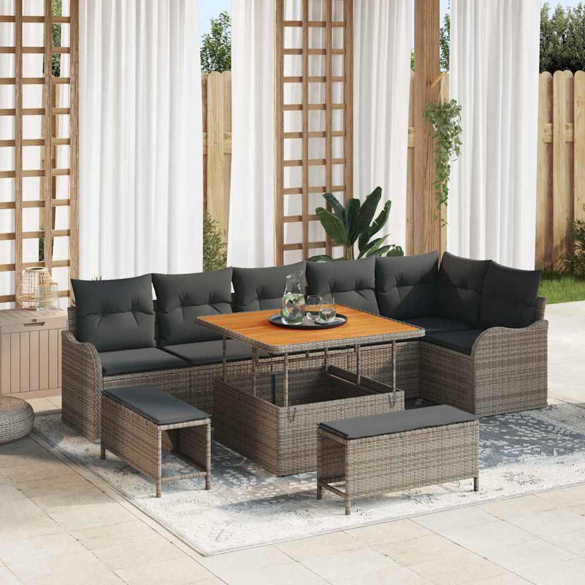 Outdoor-Ecksofa mit dunklen Kissen. Holztisch mit Getränken. Bänke. Weiße Vorhänge und Holzgitter. Grüne Pflanzen und Holzzaun.