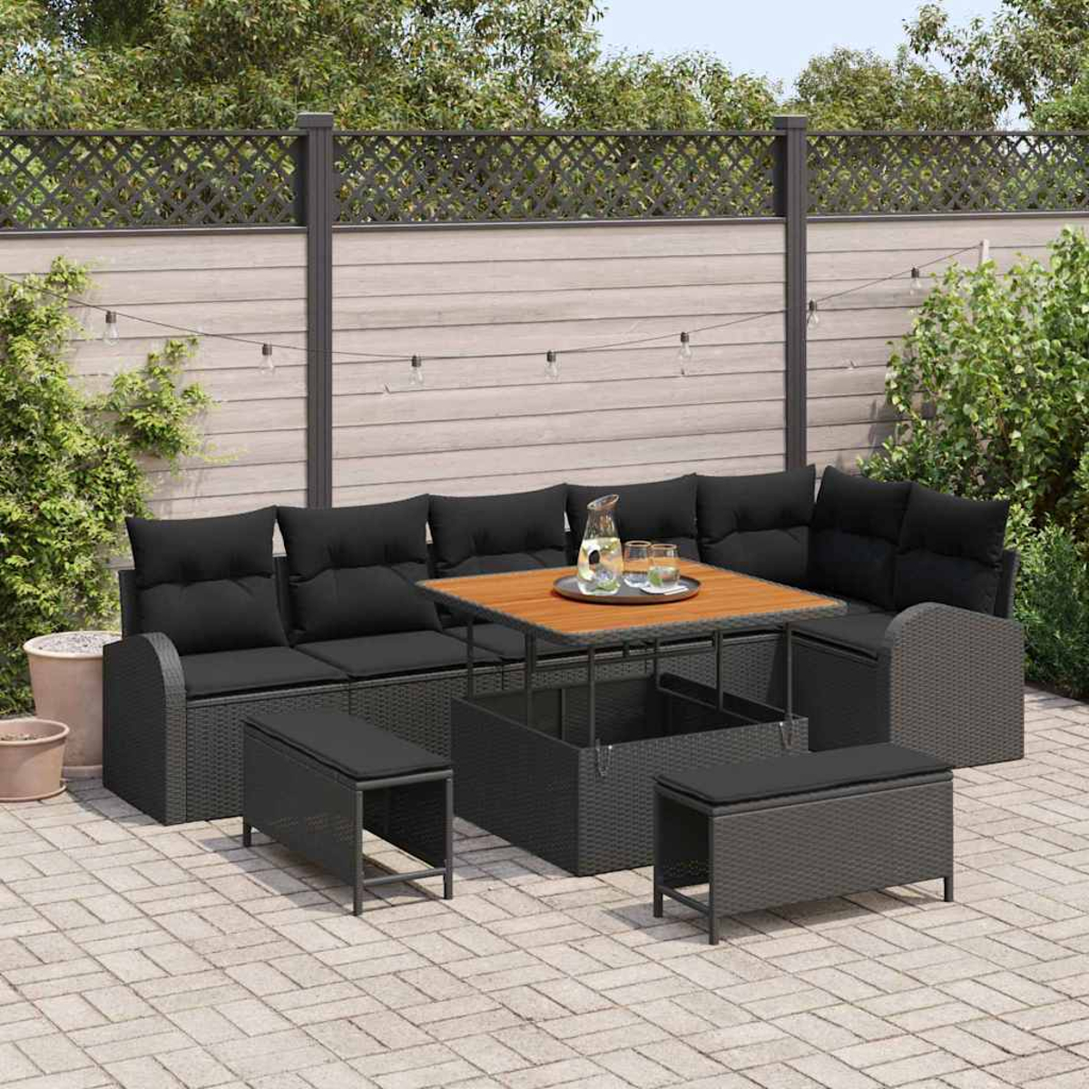 Garten-Sitzgruppe mit schwarzen Kissen, einem Holztisch und zwei Bänken. Lichterketten hängen über der Terrasse.