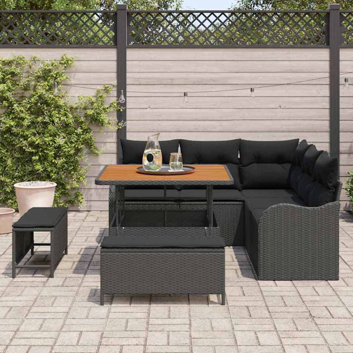 Juego de patio exterior con cojines negros y una mesa de madera. Incluye un banco y un taburete pequeño. Contra una valla de madera.