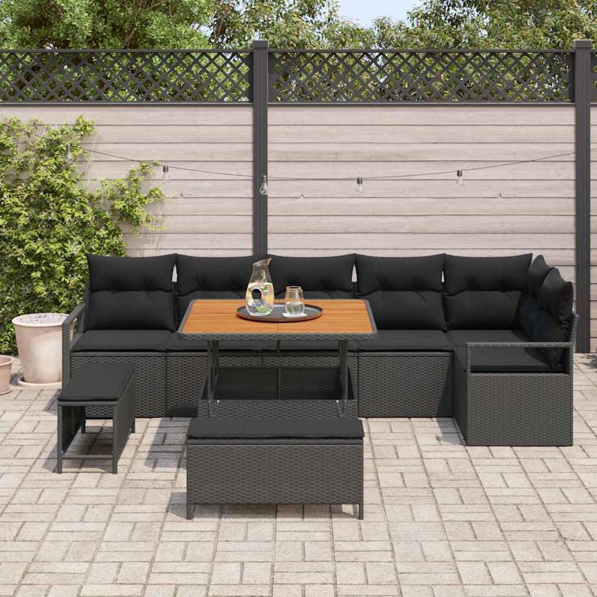 Terrassenset: graues Korbmöbel, Holztisch und zwei Ottomane. Schwarze Kissen. Vor einem Zaun und Grün.