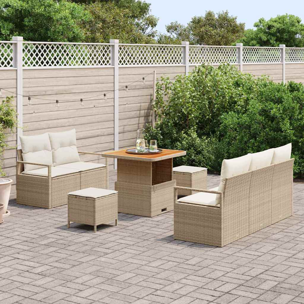 Terrassengarnitur mit gewebten beigen Möbeln, einem Tisch und Sitzgelegenheiten. Ein weißer Zaun und grüne Büsche bilden den Hintergrund.