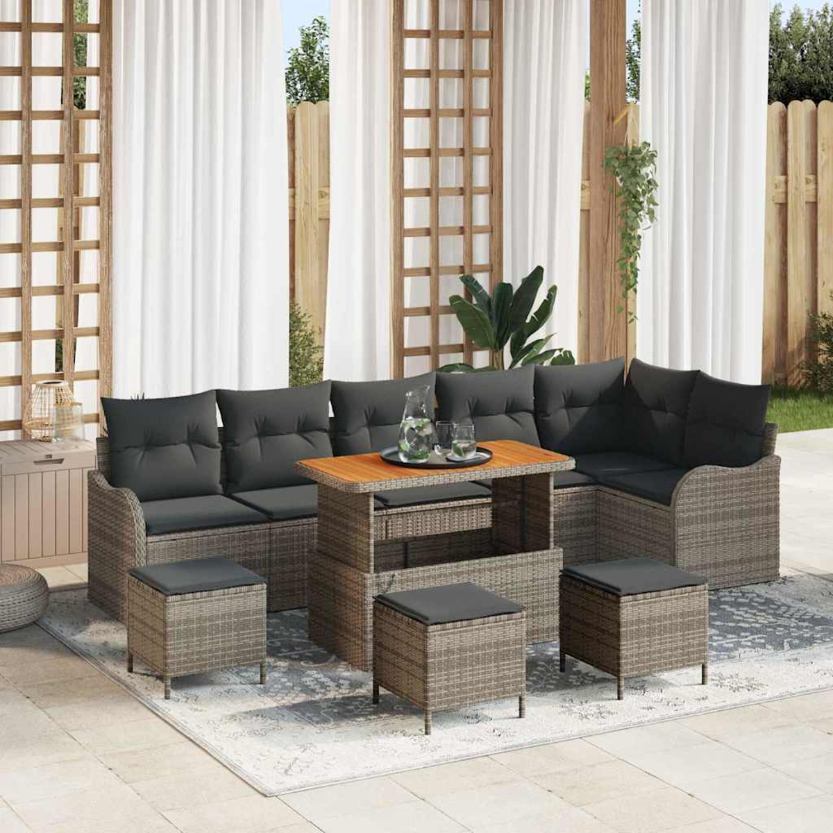 Garten-Sitzgruppe mit grauen Kissen, einem Holztisch mit Getränken und mehreren kleinen Hockern. Weiße Vorhänge und ein Holzzaun sind im Hintergrund.
