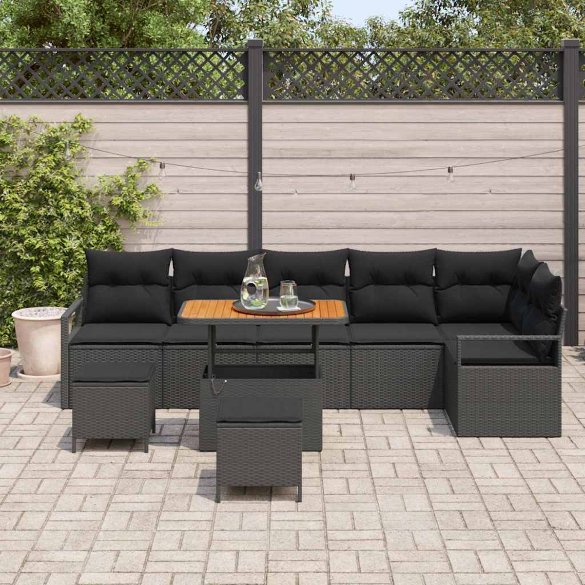 Terrassenmöbel mit schwarzen Kissen, Holztisch und grauen Korbmöbeln. Auf dem Tisch stehen eine Kanne und Gläser.