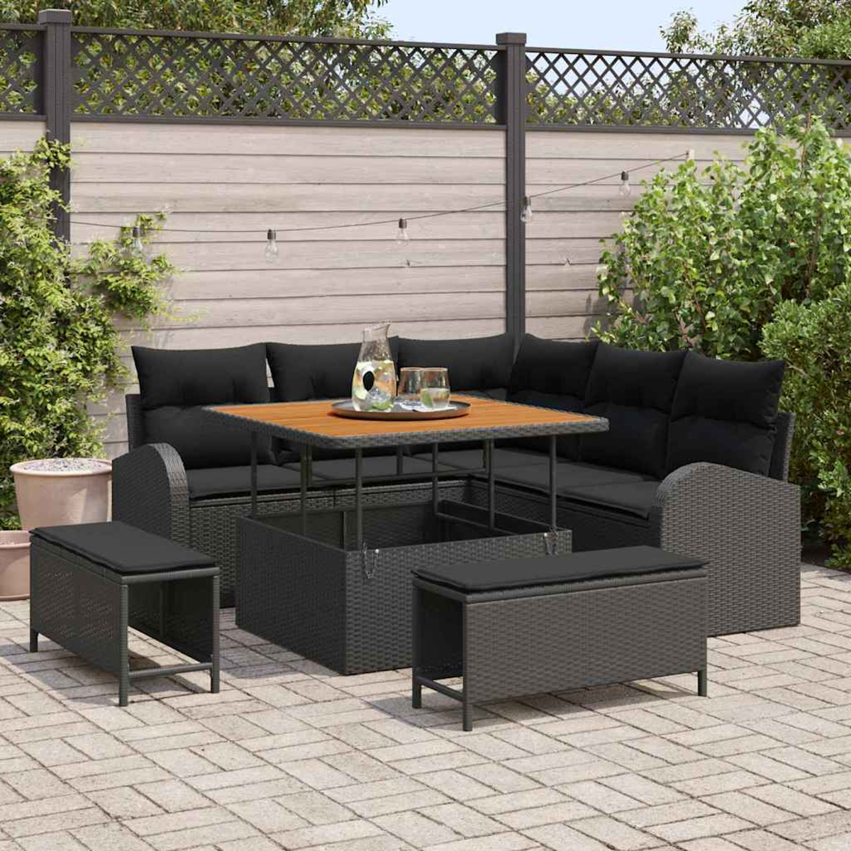 Garten-Sitzgruppe mit schwarzen Kissen, Holztisch und Bänken auf einer gepflasterten Terrasse, umgeben von Grün und Holzzaun.