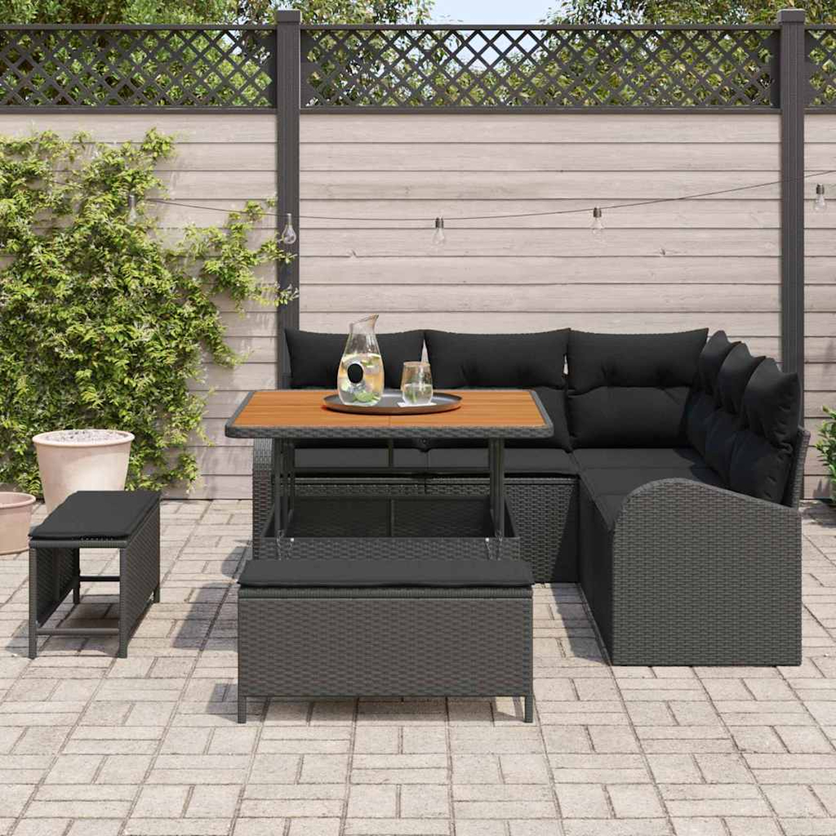 Terrassenset mit schwarzen Kissen, Holztisch und grauem Korbgeflecht. Beinhaltet Sofa, Hocker und Hocker. Hintergrund: Zaun und Pflanzen.