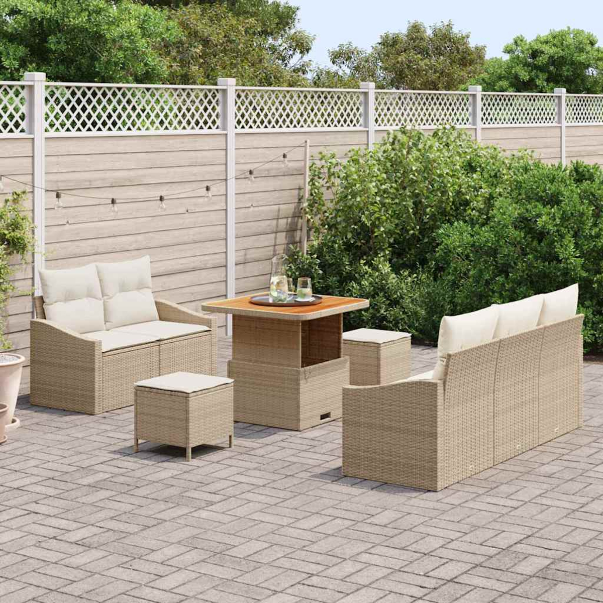 Terrassenset mit beigen Korbmöbeln, Kissen und einem Holztisch. Im Hintergrund ein weißer Gitterzaun und grüne Büsche.