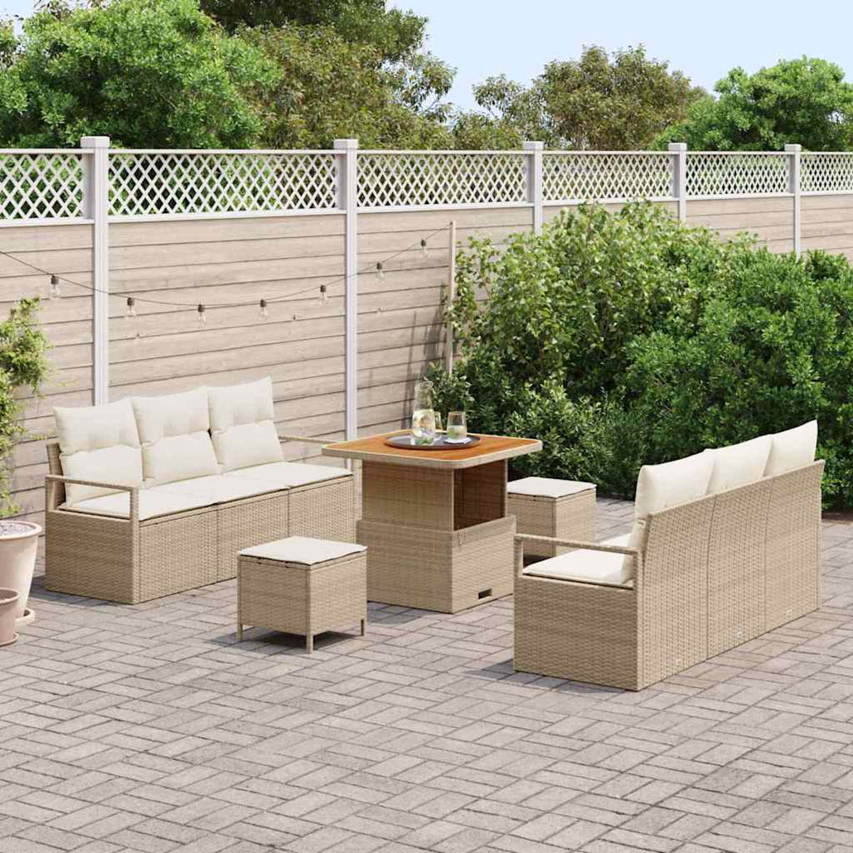 Garten-Terrassenset mit beigen Korbmöbeln, darunter Sofas, ein Tisch und Ottomanen. Im Hintergrund ein weißer Zaun und Grün.