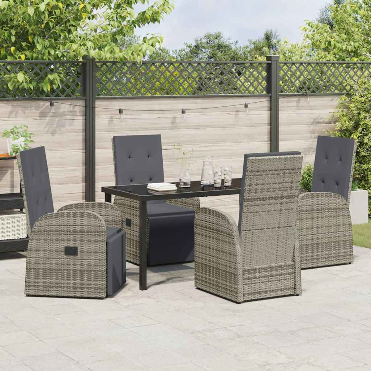 Set da patio esterno: tavolo con piano in vetro, quattro sedie intrecciate con cuscini grigi, su un patio con recinzione e verde.